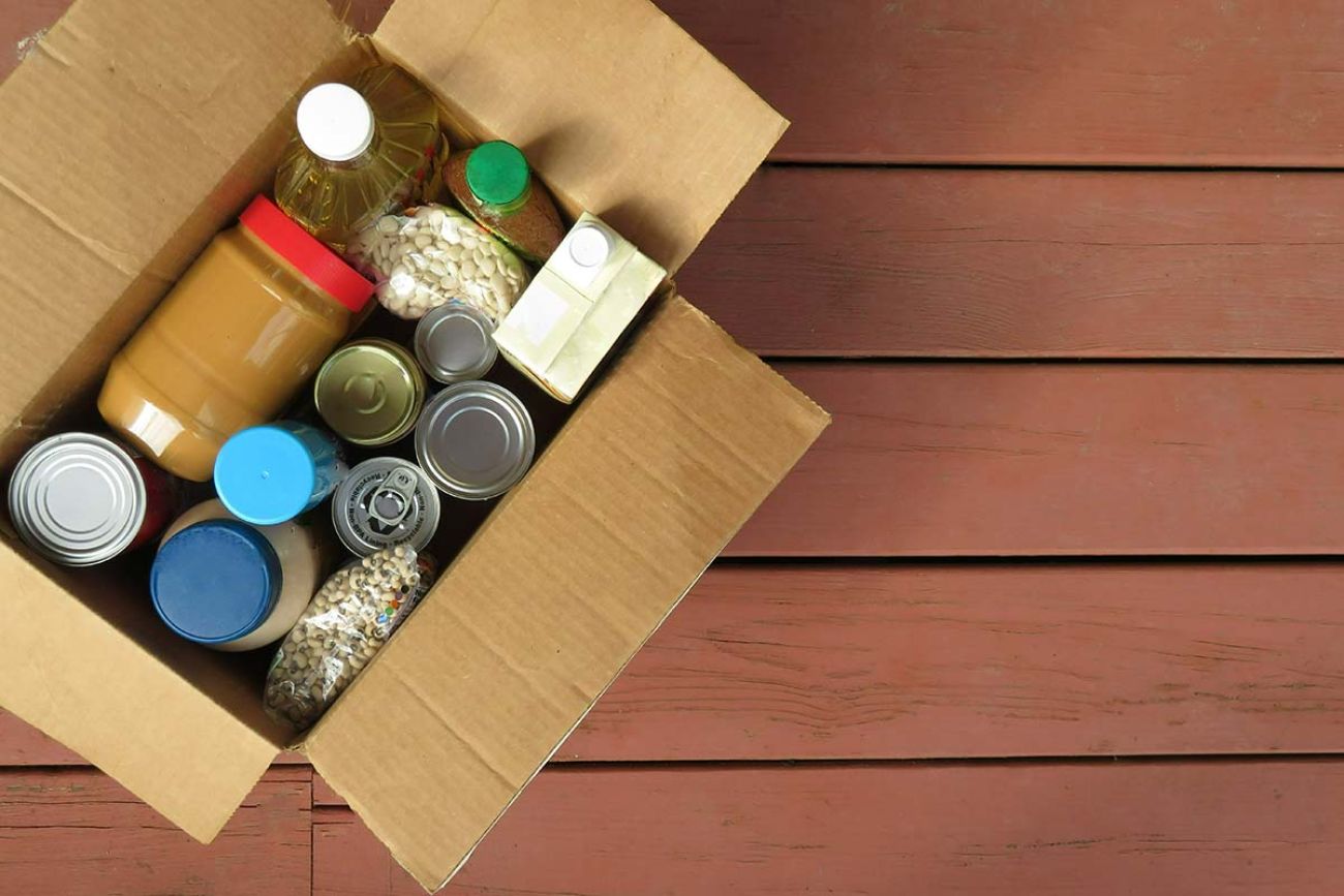 A box of donated canned goods and non-perishable foods for a food pantry for the poor sitting on red, wood panel background. 