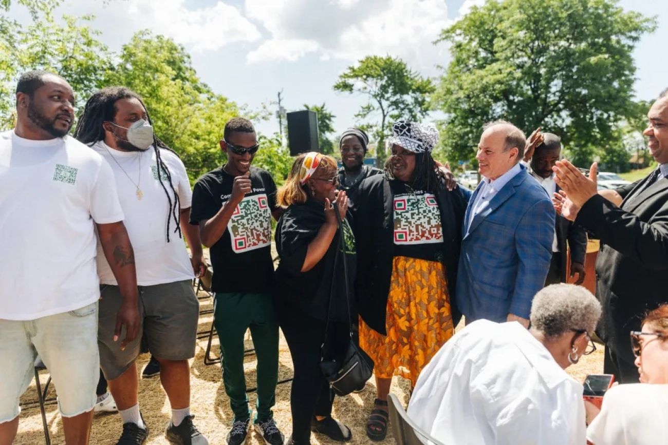 Detroit Mayor Mike Duggan, wearing a blue suit, talks to a group of residents. They are all outside