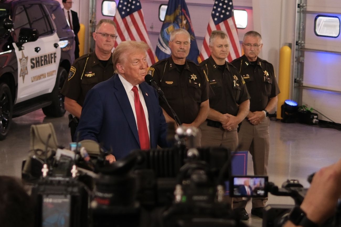 Donald Trump speaking into a microphone, law enforcement officers stand behind him