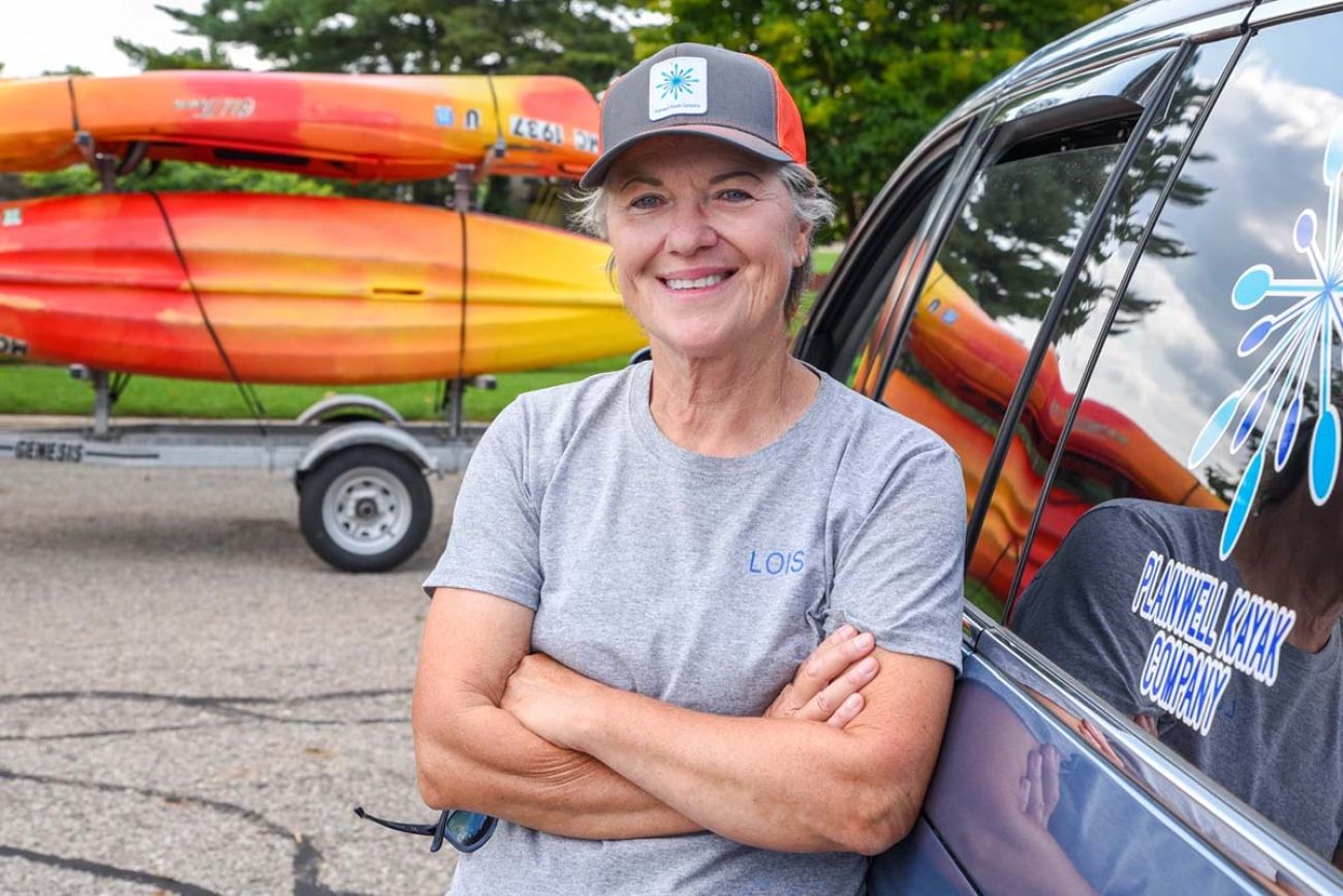 Lois Heuchert leans against a van. Kayaks are behind her