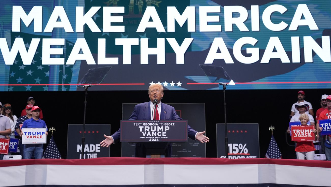 Former President Donald Trump at a rally, a sign in the back says "Make America Wealthy Again"