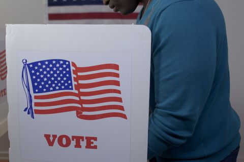 man in blue sweater and face hidden, casting vote in booth at polling station. US flag on wall in background