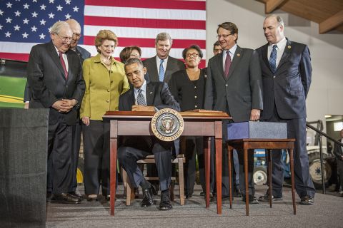 Then-President Barack Obama sitting behind a table. He is surrounded by several lawmakers. An American flag is behind the lawmakers