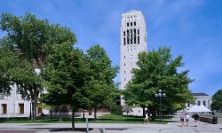 Students walk on the campus of the University of Michigan on a sunny day, with Burton Memorial Tower in the background.