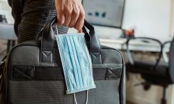 closeup of a young man in an office holding a briefcase and a surgical mask in his hand