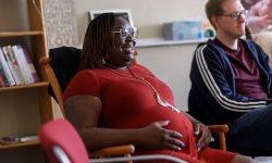 Asia LoveLady sits in a chair. She is pregnant and wearing a red dress